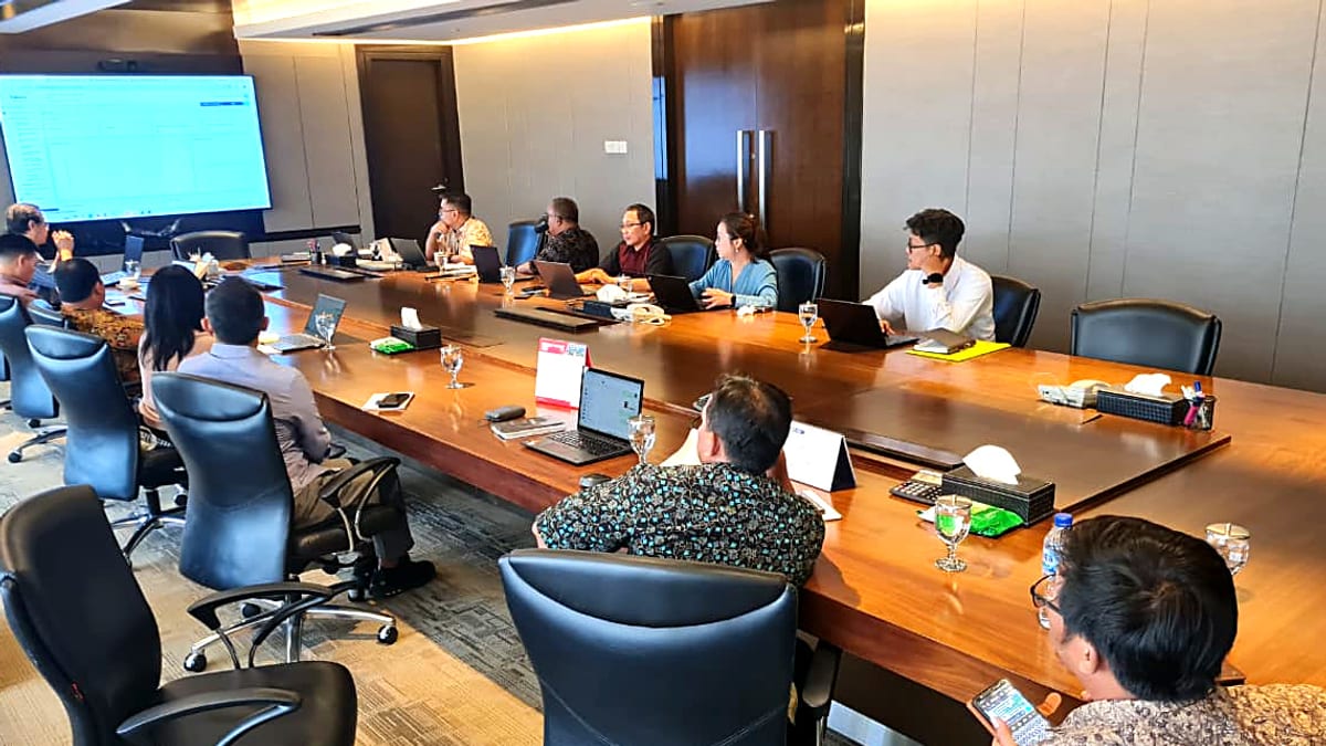 Rapat di ruang boardroom bermeja kayu besar; sekitar dua belas peserta duduk dengan laptop terbuka menghadap layar proyektor yang menampilkan aplikasi manajemen properti.